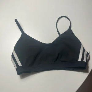 Svart Adidas linnetopp med vita ränder - Sportig svart linnetopp från Adidas med klassiska vita ränder på sidorna och justerbara axelband. Toppen har en stilren design med Adidas-logga framtill och är tillverkad i mjuk polyester som andas. Perfekt för träning eller chill dagar.