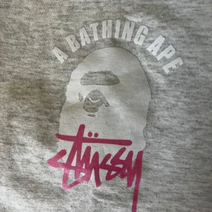 Grå BAPE x Stüssy t-shirt S - Snygg grå t-shirt från BAPE i samarbete med Stüssy. Stor tryck med BAPE-apan och Stüssy-loggan i vitt och rosa på både bröstet och ryggen. Klassisk rund hals och korta ärmar. Mjuk bomull som är skön mot huden, perfekt för streetwear. Tröjan är köpt på Myrorna och har inget tecken på användning 
