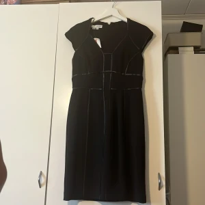 Svart kortklänning från Goddiva London - Snygg svart kortklänning från Goddiva London med coola glansiga detaljer längs sömmarna. Klänningen har en v-ringad front, korta ärmar och markerad midja som ger en stilren siluett. Perfekt för dig som gillar en modern och edgy look. Pris går att diskutera