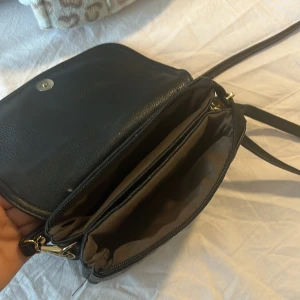 Svart quiltad axelväska Michael Kors - Snygg svart axelväska från Michael Kors med quiltad framsida och guldfärgade detaljer. Väskan har flera fack, dragkedja och magnetlås. Perfekt storlek för mobilen och det viktigaste. Stilren design med logga i metall framtill.