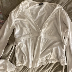 Vit omlottblus från Gina Tricot - Säljer en vit omlottblus från Gina Tricot i storlek M. Blusen har lång ärm, v-ringad hals och knytband i midjan. Materialet är lätt och skönt, perfekt för vår och sommar. Enkel och stilren design som är lätt att matcha med jeans eller kjol.