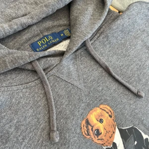 Grå hoodie Polo Bear Ralph Lauren - Säljer en grå hoodie från Polo Ralph Lauren med ikoniska Polo Bear-trycket på bröstet. Tröjan har huva med snörning och lång ärm. Materialet är mjuk bomullsmix som är perfekt för chill dagar. Klassisk och stilren look med coolt motiv.