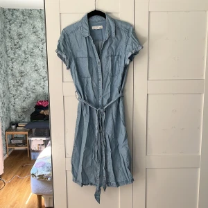 Klänning H&M - Ljusblå klänning från H&M med knappar i mitten hela vägen samt band som går att knyta i midjan. Två bröstfickor. Storlek: 38. Material: 100% lycocell. I använt skick, men inga skador eller förslitningar. 
