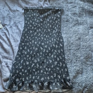 Svart blommig kortklänning Bershka - Supersöt svart tubklänning från Bershka med små vita blommor och volangkant längst ner. Klänningen är smockad och har en stretchig passform som sitter snyggt. Perfekt för dig som gillar romantiska detaljer och vill ha en klänning som sticker ut.