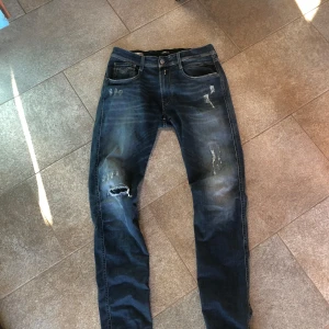 Replay Anbass blå jeans med slitningar - Säljer ett par Replay Anbass jeans i mörkblå tvätt med snygga slitningar och hål på benen. Modellen har klassisk femficksdesign, smal passform och läderpatch bak i midjan. Perfekta för dig som gillar en cool och avslappnad stil.