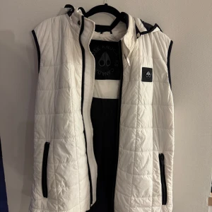 Vit dunväst Moose Knuckles - Snygg vit dunväst från Moose Knuckles med svart kant och huva som är avtagbar. Quiltad design med två svarta dragkedjefickor och logopatch på bröstet. Västen är gjord i polyester och har en clean, sportig look som funkar till streetwear. Perfekt för lager-på-lager under kyliga dagar. Inte använd mycket alls! Väldigt bra skick.