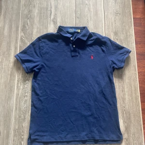 Marinblå Polo Ralph Lauren piké - Snygg marinblå piké från Polo Ralph Lauren med klassisk pikékrage och två knappar framtill. Custom slim fit-modell med röd broderad logga på bröstet. Tillverkad i mjuk bomull som känns skön mot huden. Perfekt för dig som gillar stilren och enkel design.