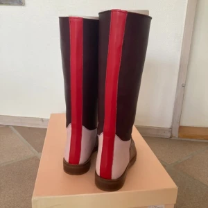 SÖKES! Stine goya stövlar - stl 39 - Säljer ett par höga stövlar med cool färgblock-design i brunt, vinrött och ljusrosa, med en röd rand längs hela skaftet. Stövlarna har platt sula och är tillverkade i skinn, vilket ger dem en lyxig känsla och snygg finish. Perfekt för dig som gillar att sticka ut med din stil.