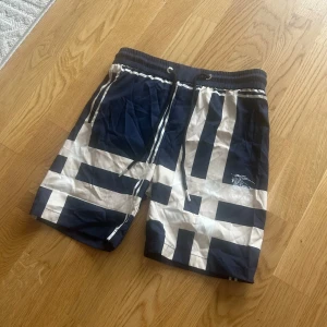 Blå Burberry shorts  - Snygga shorts från Burberry i marinblått och vitt med grafiskt rutigt mönster. De har elastisk midja med snörning och Burberry-logga på framsidan. Materialet är lätt och torkar snabbt, perfekt för sommardagar vid stranden eller poolen.