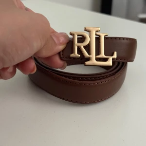 Brunt skärp från Ralph Lauren RL - Snyggt brunt bälte från Ralph Lauren med ett stort RL-spänne i guld. Bältet är gjort i slätt skinn och har stilrena sömmar längs kanterna. Perfekt accessoar för att lyfta din outfit med en lyxig touch.