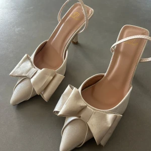 Beige klackskor  med rosett från Givana - Säljer mina snygga beige pumps från Givana med spetsig tå och stor rosett framtill. Skorna har en smal rem runt hälen och låg klack. Materialet har en lätt satin-finish som ger en lyxig känsla. Perfekta för dig som gillar eleganta detaljer . Skorna är knappt använda och skickar även med kartongen