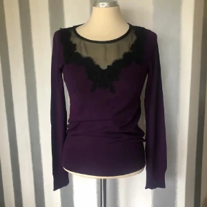 Lila topp med spetsdetalj - Snygg lila långärmad topp med transparent mesh och svart spets framtill vid halsen. Figurnära passform och mjukt material som känns skönt mot huden. Perfekt för dig som gillar unika detaljer och vill sticka ut lite extra.