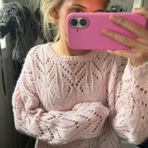 Ljusrosa stickad tröja med mönster - Superfin ljusrosa stickad tröja med ett snyggt hålmönster och rund hals. Tröjan har långärmade, lite loose ärmar och känns riktigt mjuk och fluffig. Perfekt att styla med jeans eller kjol för en chill look.