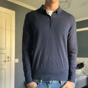 Mörkblå stickad pikétröja från Uniqlo - Snygg mörkblå pikétröja från Uniqlo i merinoull Tröjan har klassisk krage, två knappar framtill och långa ärmar. Perfekt för dig som gillar stilrena och enkla plagg med en chill vibe. Passar dig som vill ha en normal passform och diskret look.         På sista bilden kan man se att jag har sytt igen ett hål i ena armhålan på tröjan vilket inte syns vid användning.
