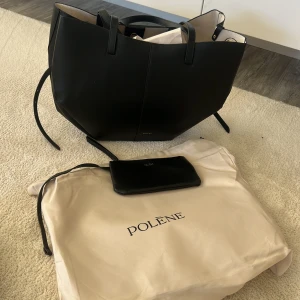 Svart shoppingväska från Polène - Stilren och stor svart shoppingväska från Polène i skinn med minimalistisk design och två rejäla handtag. Väskan har en mjuk beige insida och kommer med en matchande svart plånbok. Perfekt för dig som gillar snygga och praktiska accessoarer. Fick den i present men säljer vidare då den inte var så som jag tänkt 
