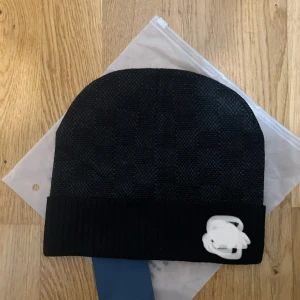 Svart rutig mössa  - Snygg svart mössa med diskret rutigt mönster och ribbad kant. Märkets ikoniska logga i vitt sitter på kanten och ger en lyxig touch. Perfekt för dig som vill ha en stilren och exklusiv look.