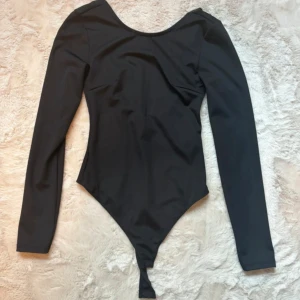 Svart långärmad bodysuit med djup rygg - Snygg svart bodysuit med lång ärm och djup, rundad rygg. Kroppsnära passform och stretchigt material som ger en clean och stilren look. Perfekt att styla med jeans eller kjol för en trendig outfit.