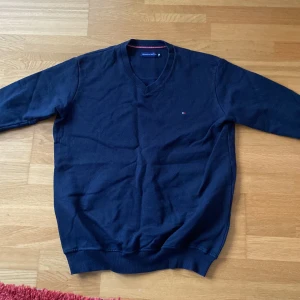 Marinblå v-ringad tröja Tommy Hilfiger - Snygg marinblå v-ringad tröja från Tommy Hilfiger med diskret logga broderad på bröstet. Tröjan har långa ärmar och ribbade muddar vid hals, ärmslut och nederkant. Tillverkad i mjuk bomull som är skön mot huden. Perfekt för dig som gillar klassisk stil med en modern touch.