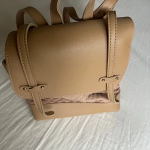 Beige ryggsäck i syntetmaterial - Säljer en stilren beige ryggsäck i läder  med quiltad detalj på framsidan. Väskan har justerbara axelremmar och ett lock med dekorativa sömmar och metalldetaljer. Perfekt för dig som vill ha en snygg och praktisk accessoar.
