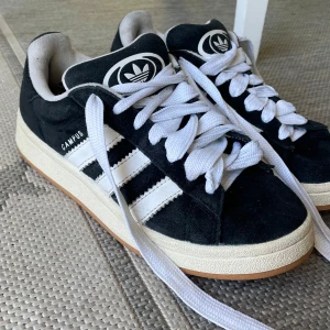 Svarta Adidas Campus sneakers - Säljer ett par klassiska Adidas Campus00s sneakers i svart mocka med vita ränder och vit sula. Skorna har breda vita skosnören och gummisula i beige. Perfekta för dig som gillar retrostil och streetwear. Bra skick förutom att de har två små håll inuti skon på hällarna , se sista bild!!!! Och lite slitning på baksidan av skon längst ner💞men jätte fina o tvättat rent dom så dom är helt rena o fina. Ny pris: ca 1200kr , har tyvärr inte kvar original kartongen. Men kan skicka äkte bevis.