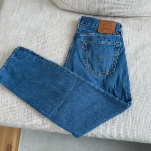 Blå Levis 501 jeans - Klassiska blå Levis 501 jeans med rak passform och femficksdesign. Byxorna har en brun läderpatch bak i midjan och synlig röd Levis-tagg på bakfickan. Perfekta för en tidlös och avslappnad stil.