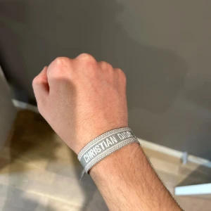 Dior armband  - Säljer ett stilrent grått armband från Dior med tydlig logga i vitt. Armbandet är vävt och har en enkel, modern design som passar perfekt till din accessoarsamling.