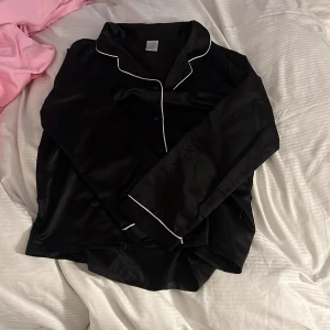 Svart satinpyjamas från Ladies Lounge - Säljer en svart långärmad pyjamasskjorta i satin från Ladies Lounge. Skjortan har vita kontrastkanter och knäppning framtill. Perfekt för mysiga hemmakvällar eller som loungewear.