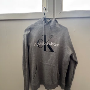 Calvin Klein Hoodie – Grå M (Unisex Fit, Logga fram) - Stilren hoodie från Calvin Klein Jeans i klassisk grå färg med stor CK-logga framtill!