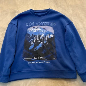 Blå sweatshirt med Los Angeles-tryck - Säljer en blå sweatshirt med tryck av snötäckta berg och texten 'Los Angeles' framtill. Tröjan har rund hals och långa ärmar, perfekt för en avslappnad stil.