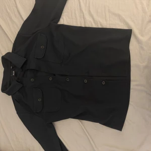 Svart overshirt med fickor - Säljer en svart overshirt från H&M med två stora bröstfickor och knäppning framtill. Skjortan har klassisk krage och långa ärmar, 