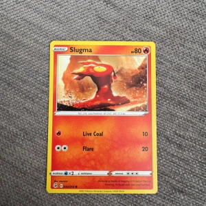 Pokémonkort Slugma - Säljer ett Pokémonkort med Slugma, en eldstyp med 80 HP. Kortet har attacker som Live Coal och Flare. Framsidan har en bild på Slugma i ett eldigt landskap och baksidan har klassisk Pokémon-design med Pokéboll.