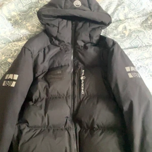 Svart dunjacka från Sail Racing - Säljer en svart dunjacka från Sail Racing med broderad logga på bröstet och ärmarna. Jackan har huva och dragkedja framtill samt patch med texten 'Sail Antarctica Expedition'. Perfekt för kalla dagar.