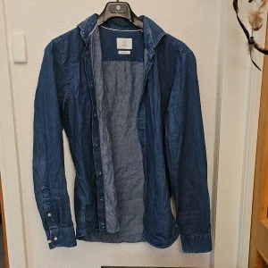 Mörkblå jeansskjorta från Bläck - Snygg mörkblå jeansskjorta från Bläck med klassisk krage och knappar framtill. Skjortan har långa ärmar med manschett och är tillverkad i ett mjukt denimtyg. Perfekt för en avslappnad men stilren look.