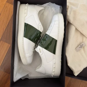 Valentino Garavani Open | ev byte - Säljer ett par vita Valentino Garavani Open sneakers med en bred grön lackad rem över mitten. Skorna har vita snören, rund tå och små nitar på hälen. Använda max 3 gg kan även bytes