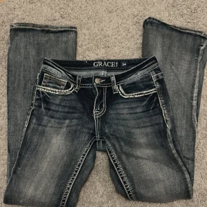 Low waist bootcut jeans med broderade bakfickor - Snygga mörkblå jeans med låg midja från Grace in LA med slitningar och kontrastsömmar. Bakfickorna har broderade detaljer och små blommor. Klassisk femficksmodell med knapp och dragkedja. Perfekta för dig som gillar jeans med lite extra detaljer. Storlek 14 men skulle säga att de passar folk med storlek 152. Säljer då de är försmå. Köpts för 900kr
