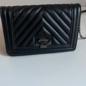 Svart axelväska - Snygg svart axelväska med quiltat mönster och silvrigt spänne framtill. Väskan har en kedjerem och ett innerfack med dragkedja. Perfekt storlek för det viktigaste och passar till många olika outfits.