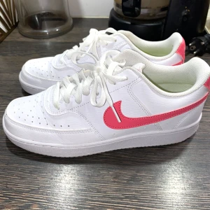 Nike sneakers  - Säljer ett par fräscha vita Nike sneakers med rosa swoosh och häl. Skorna har klassisk design med snörning, perforerad tå och platt sula. Perfekta för dig som gillar sportig och stilren look. Använt endast 1 gång. 