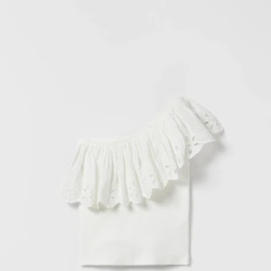 Vit oneshoulder topp med volang - Säljer en vit oneshoulder topp med bred volangkant och broderade detaljer. Perfekt för sommardagar och ger en romantisk känsla till outfiten. Tunn och luftig modell.