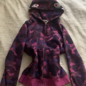 Lila camo zip hoodie från BAPE - Säljer en lila och rosa camo zip hoodie från BAPE med hajmotiv och broderade bokstäver på huvan. Jackan har dragkedja hela vägen, dock är dragkedjan lite krånglig ibland upp och fickor framtill. Knappt använd bara legat i garderoben ett tag. Bra skick💕strl m fast passar mig som har xs/s lite baggy. 