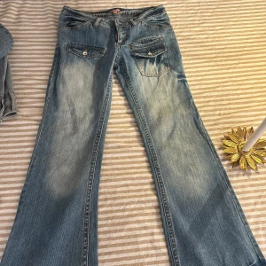 Blå bootcut jeans med fickdetaljer - Säljer ett par blå bootcut jeans med slitningar och snygga fickor med lock och knappar både fram och bak. Jeansen har låg midja och klassisk femficksdesign. Perfekta för dig som gillar en retroinspirerad look. Säljer då de är för små på mig