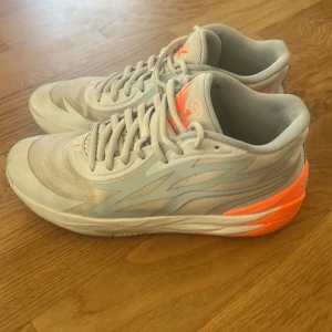 Gråa Lamello ball skor från puma  - Säljer ett par grå sneakers med ljusblå detaljer och starkt orange häl från MB1. Skorna har snörning, rund tå och en platt sula. De har en sportig design med tryckt vinge och nummerdetaljer på plös och häl.