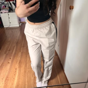 Beiga träningsbyxor med svarta detaljer - Säljer ett par beiga träningsbyxor med svarta sidopaneler och elastisk midja med dragsko. Byxorna är tillverkade i en blandning av polyester och nylon, vilket gör dem lätta och bekväma. Perfekta för en aktiv vardag.