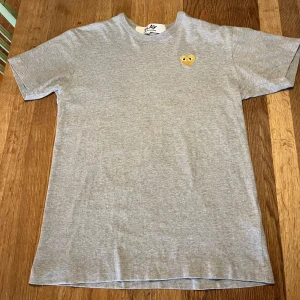 Grå t-shirt från Comme des Garçons Play - Säljer en grå t-shirt från Comme des Garçons Play med det ikoniska broderade hjärtat i guld på bröstet. T-shirten har klassisk passform och är tillverkad i mjuk bomull. Perfekt för en stilren och avslappnad look.