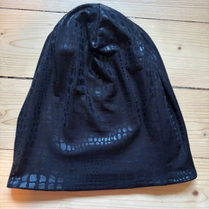 Svart mössa med glansigt ormskinnsmönster - Cool svart mössa med glansigt ormskinnsmönster. Tunn och mjuk, perfekt för att ge din outfit en edgy touch. Passar både tjejer och killar.