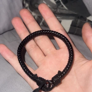 Svart flätat armband med justerbar knut - Säljer ett svart armband med flätad design och justerbar knut. Armbandet är gjort av syntetmaterial och passar perfekt som en enkel accessoar till vardagslooken. Armbandet kom i samband med en klocka men armbandet var tyvärr för stort