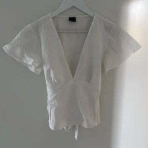 Vit omlottopp med knytband - Säljer en vit omlottopp med djup v-ringning och korta ärmar från Gina tricot. Toppen har knytband i midjan som knyts i ryggen för en snygg passform. Perfekt till sommaren och andvänd enbart en gång ❤️
