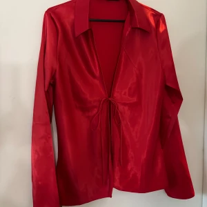 Röd satinblus med knyt - Snygg röd blus i glansigt satinmaterial med lång ärm och knyt framtill. Djup v-ringning och klassisk krage ger en trendig look. Perfekt för dig som vill sticka ut med färg och stil.