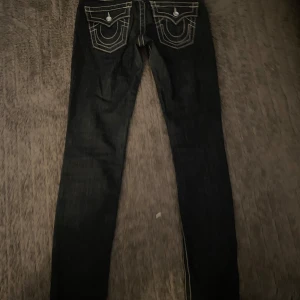 True Religion skinny jeans med kontrastsömmar - Mörkblå skinny jeans från True Religion med tydliga vita kontrastsömmar och dekorativa bakfickor. Klassisk femficksmodell och låg midja. Snygga detaljer och ikonisk etikett bak. Low waist