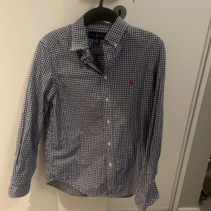 Blårutig skjorta från Ralph Lauren - Klassisk blårutig skjorta från Ralph Lauren med knappar framtill och button-down krage. Skjortan har lång ärm och en liten broderad logga på bröstet. Perfekt för en stilren och tidlös look.
