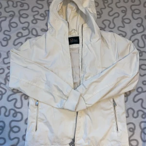 Vit vindjacka från Peak Performance - Snygg vit vindjacka från Peak Performance med huva och dragkedja framtill. Jackan har två fickor med dragkedja och är tillverkad i polyester och bomull. Jag är lite smutsig men går att tvätta bort utan problem.